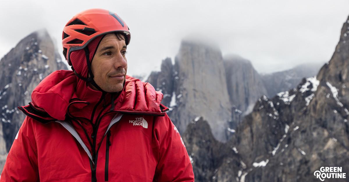 Alex Honnold wears an orange helmet and red jacket with snowy mountains in the distance