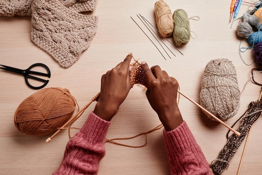 A person knitting at a table with skeins of yarn and knitting supples around the. 