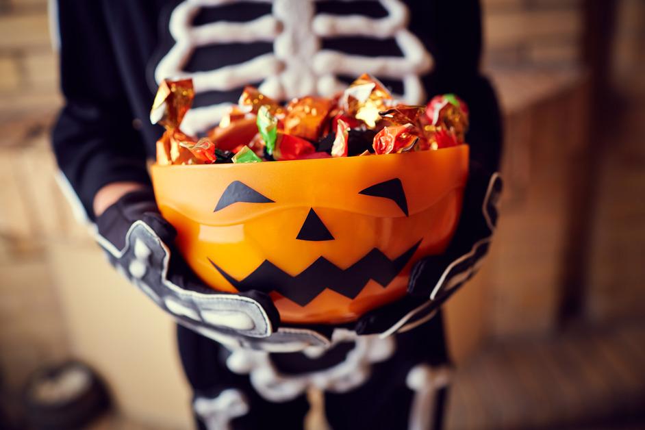 A person wearing a skeleton suit holding an orange bowl with a jack-o-lantern face full of candy. 