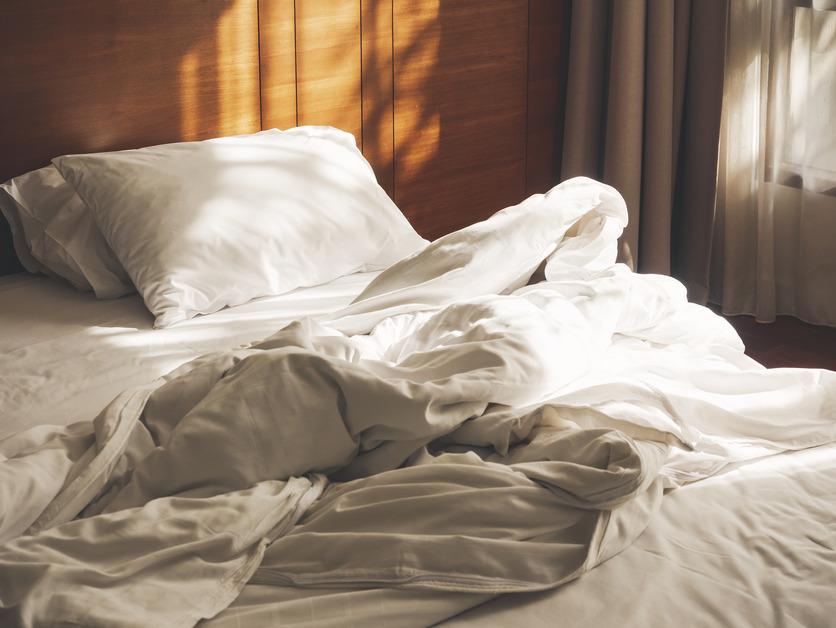 An unmade bed with white sheets and a white duvet and pillow in the morning sunlight. 
