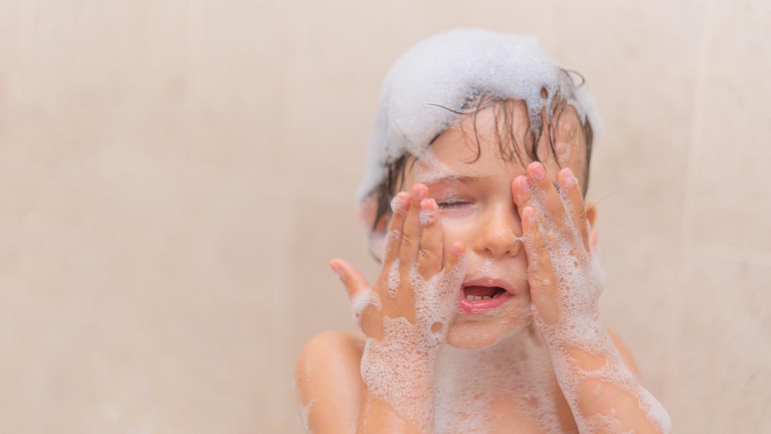 A little boy takes a bath and rubs soap out of his eyes. 