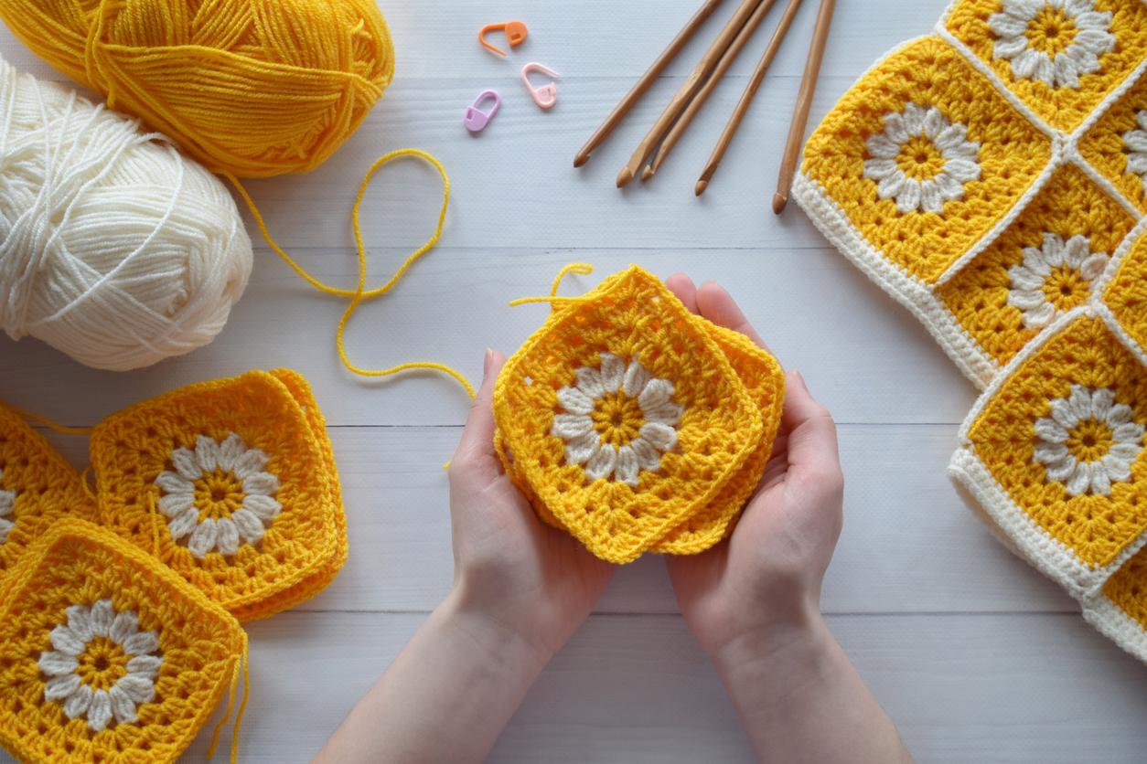 Person holding a few yellow granny squares with white flowers on them while surrounded by other crochet supplies.