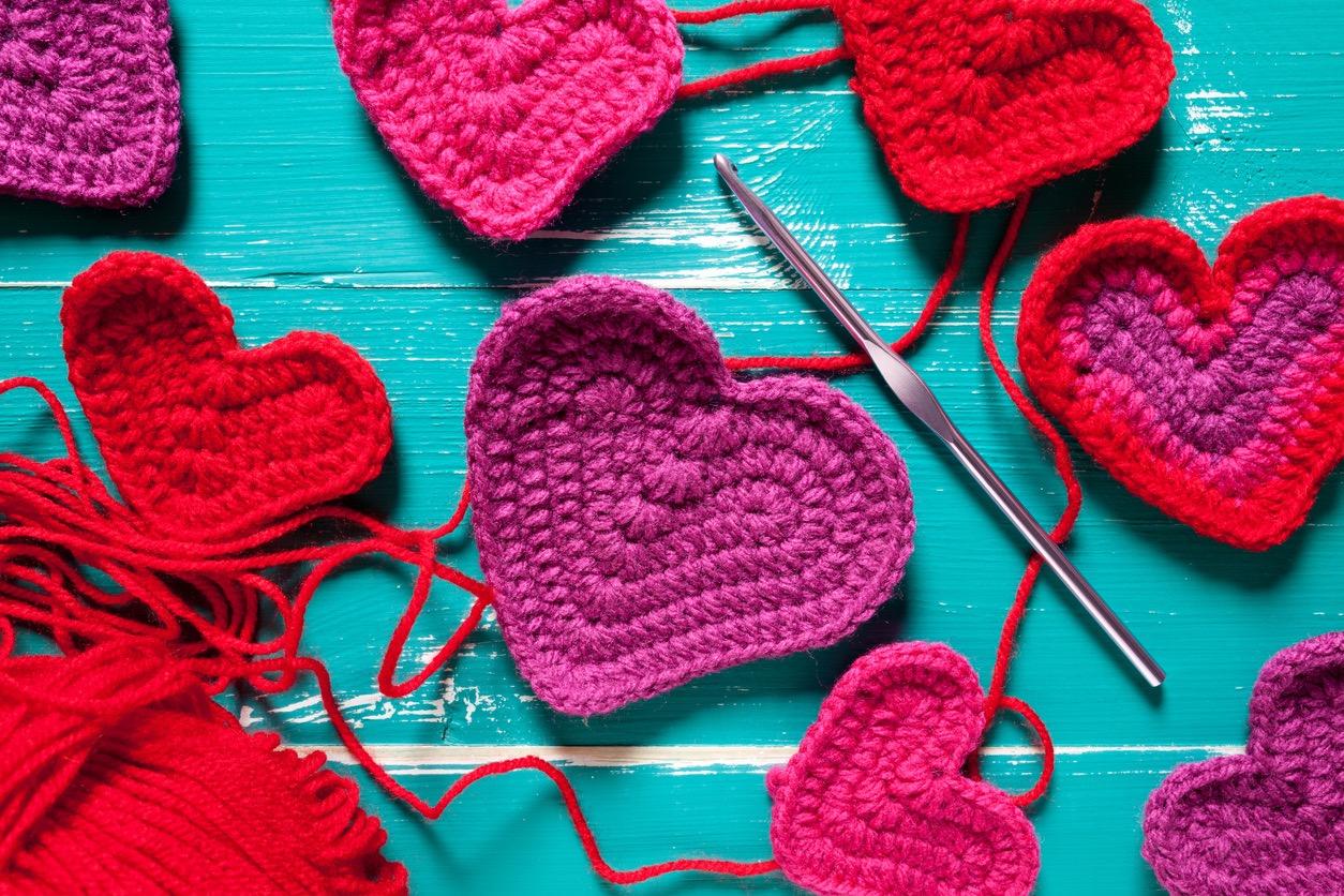 Close up of various sized purple and red crochet hearts scattered on a blue table.