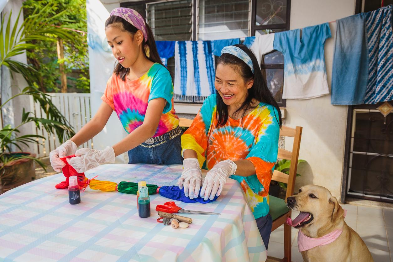 A mother and daughter tie dye shirts together on the patio beside their smiling dog.