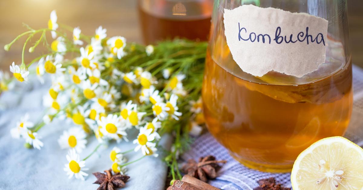 A jar with kombucha in it, with "kombucha" written on the jar, beside a bouquet of daisies and a lemon wedge.