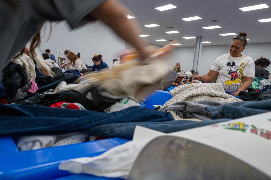 A low angle photo of people digging through blue bins at a Goodwill Outlet store, a blurry item of clothing flies across the top. 
