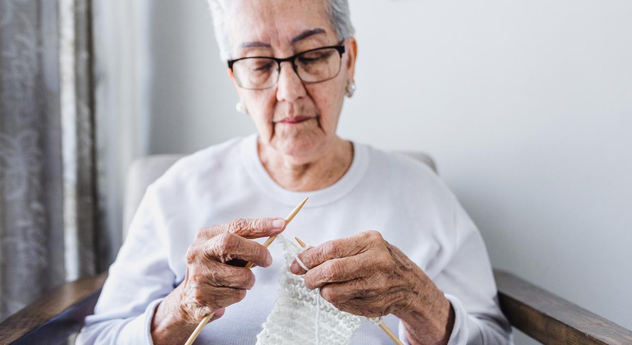 Person holding two knitting needles and working on a project made of white yarn while sitting down.