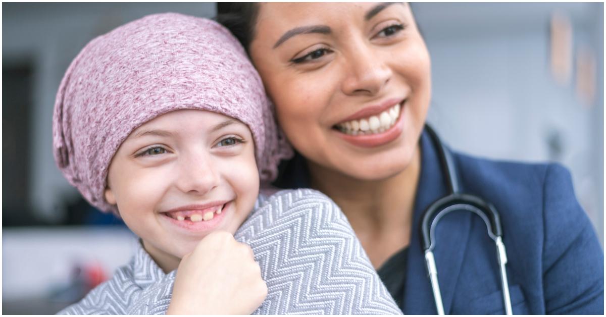 A young sick girl with a pink head covering smiles next to a female doctor. 