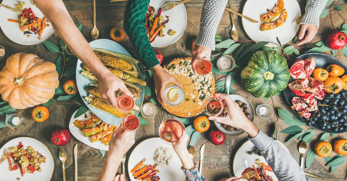 Over a dinner table covered in plates of food, various hands hold glasses in a toast, from a top view.