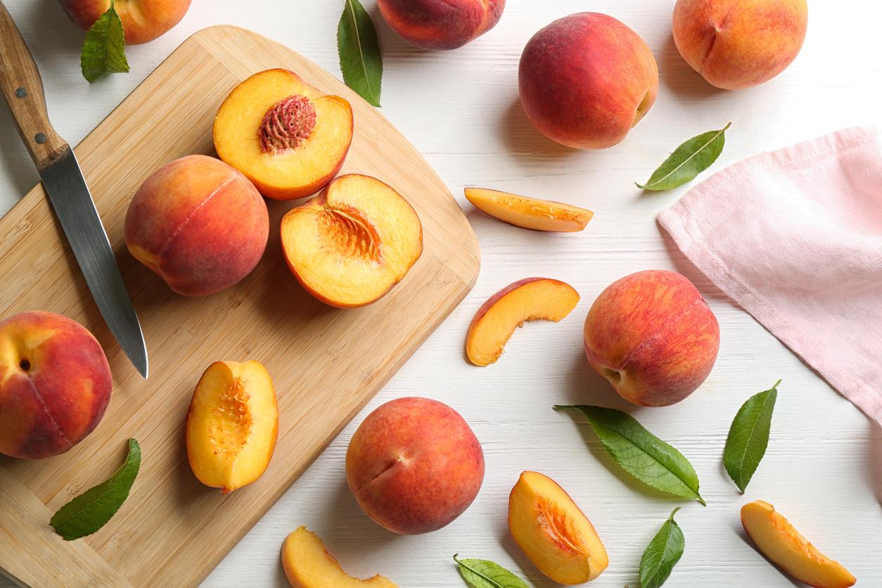 A group of peaches are cut open on a cutting board, laying beside whole peaches and green leaves on a white table cloth.