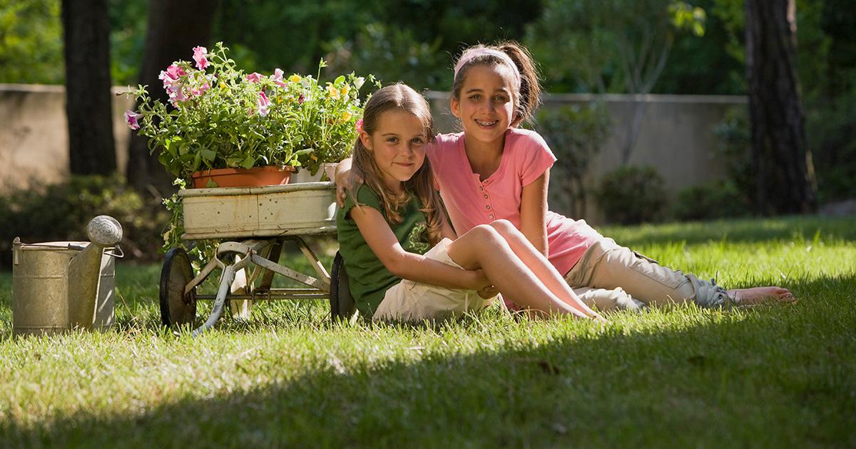 Two young girls sit in a partially shaded yard with flowers ready to be planted. 