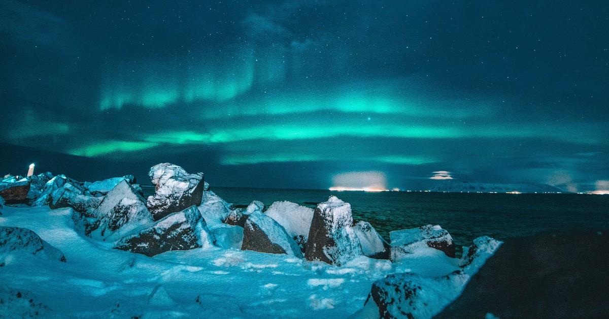The Northern Lights can be seen over the water as snow covered rocks sit in the foreground 