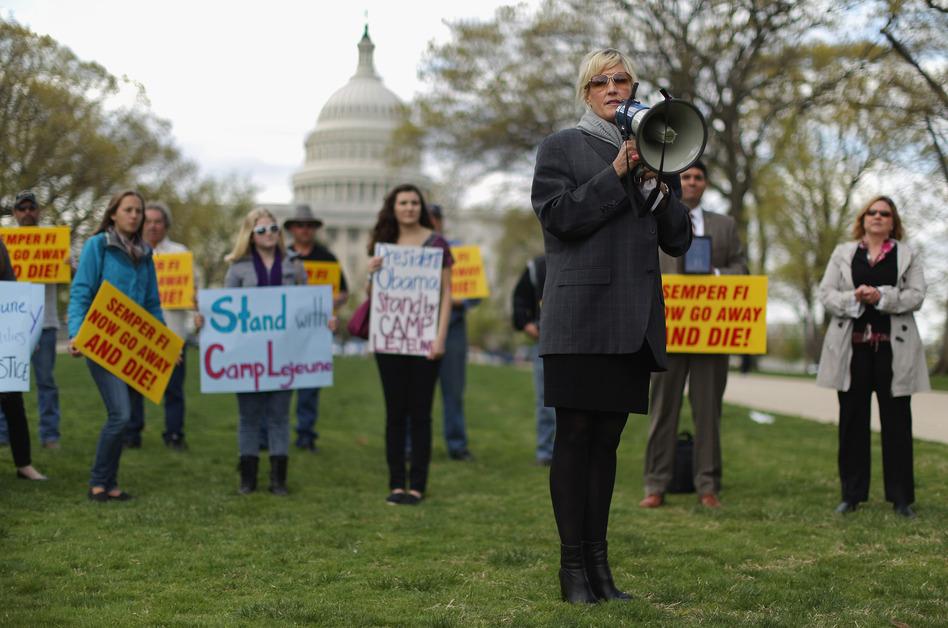 Woman with a megaphone stands in front of protestors in front of the White House with signs that say "Stand with Camp Lejeune."
